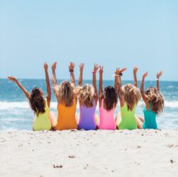 back view photo of six girls wearing swimsuit sitting on white sand