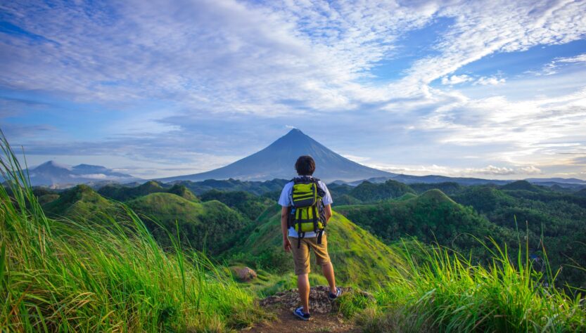 man wearing white shirt brown shorts and green backpack standing on hill
